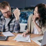 A couple reviews bills and documents on a laptop while discussing household finances.
