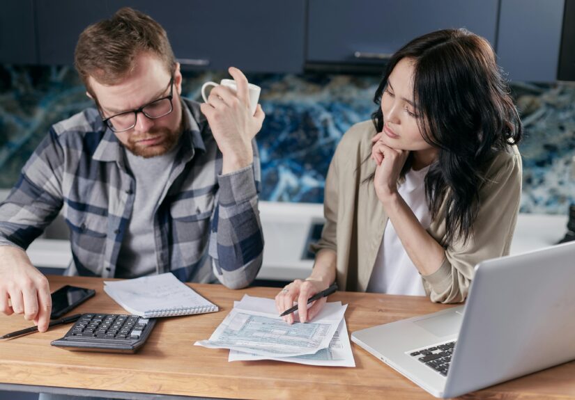 A couple reviews bills and documents on a laptop while discussing household finances.